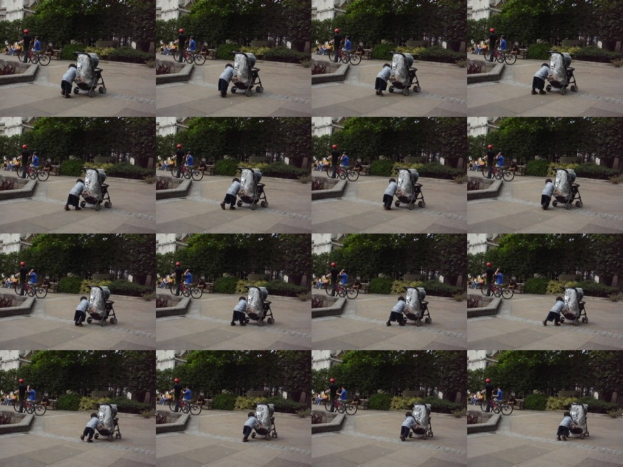 Ein Kind geht auf einer Straße und zieht einen Karren, mit zwei Personen auf der linken Seite, die Fahrräder halten, Bäume im Hintergrund und ein Gebäude auf der linken Seite.