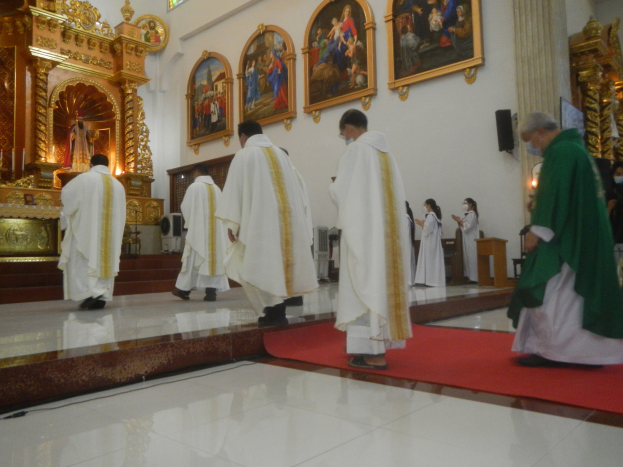Eine Gruppe von Priestern geht einen roten Teppich in einer Kirche entlang, während Menschen auf dem Boden vor ihnen und auf der rechten Seite stehen und Fotorahmen an der Wand im Hintergrund zu sehen sind.