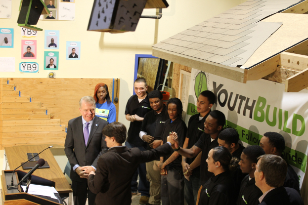 Gruppe von Menschen, die sich die Hände schütteln, um einen Podium mit Mikrofonen und Papieren auf einem Tisch, vor einer Menge, mit Plakaten an der Wand und Gegenständen auf dem Dach und der rechten Seite.