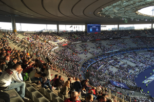 Großes Publikum in einem Stadion bei einem Fußballspiel mit einer Bühne rechts, Fahnen, Stangen, einem Bildschirm und der Allianz Arena in München, Deutschland im Hintergrund.