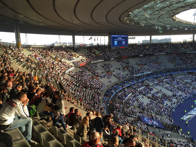 Großes Publikum in einem Stadion bei einem Fußballspiel mit einer Bühne rechts, Fahnen, Stangen, einem Bildschirm und der Allianz Arena in München, Deutschland im Hintergrund.