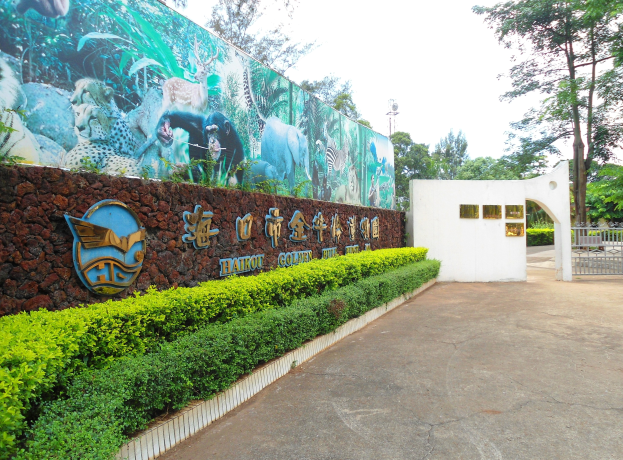 Eintritt zu einem Zoo mit einem großen Wandgemälde, das Tiere und Text zeigt, einem Tor, einem Laternenmast, umgeben von Pflanzen, einer Wand, Bäumen und einem klaren blauen Himmel.