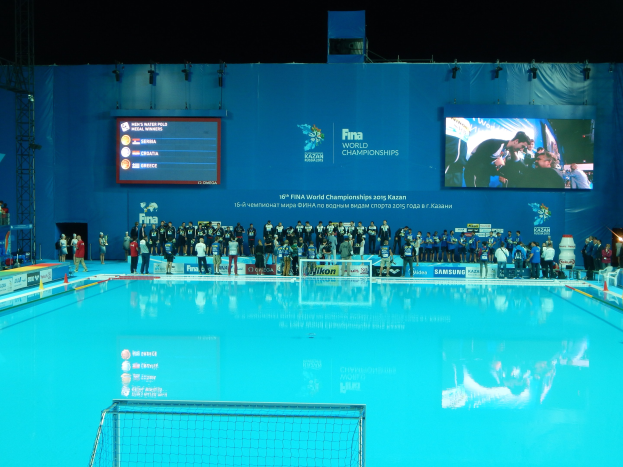 Eine Gruppe von Menschen um einen Swimmingpool mit Netz am Boden herumstehend, mit einem Bildschirm im Hintergrund, der "FINA World Championships 2015 Kazan" anzeigt.