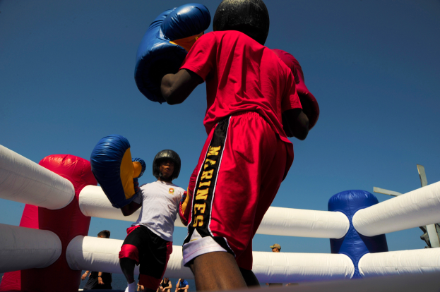 Zwei Boxer in einem Boxring, einer in rotem Hemd und blauen Shorts, kämpfen mit Boxhandschuhen, umgeben von ein paar Menschen und unter einem klaren blauen Himmel.
