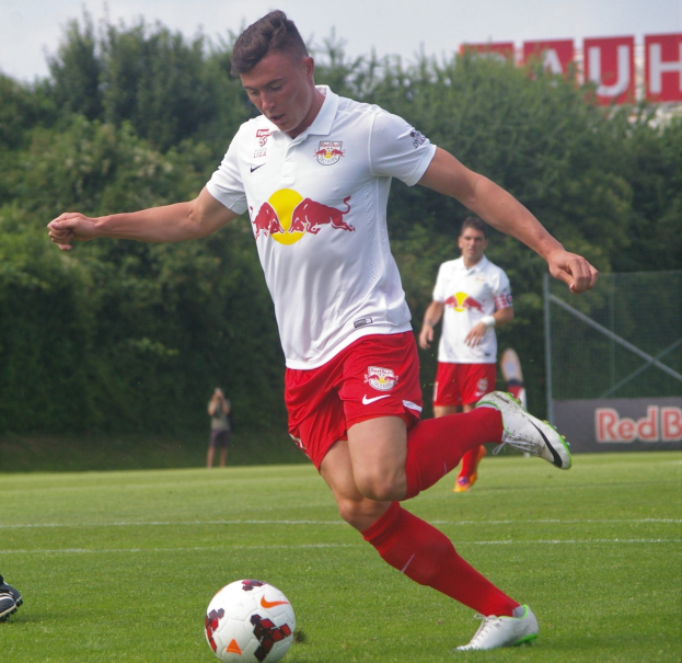 Ein professioneller Fußballspieler kickt einen Ball auf einem grünen Feld mit Bäumen und einem klaren blauen Himmel im Hintergrund, mit «RB Leipzig» auf einer Tafel.