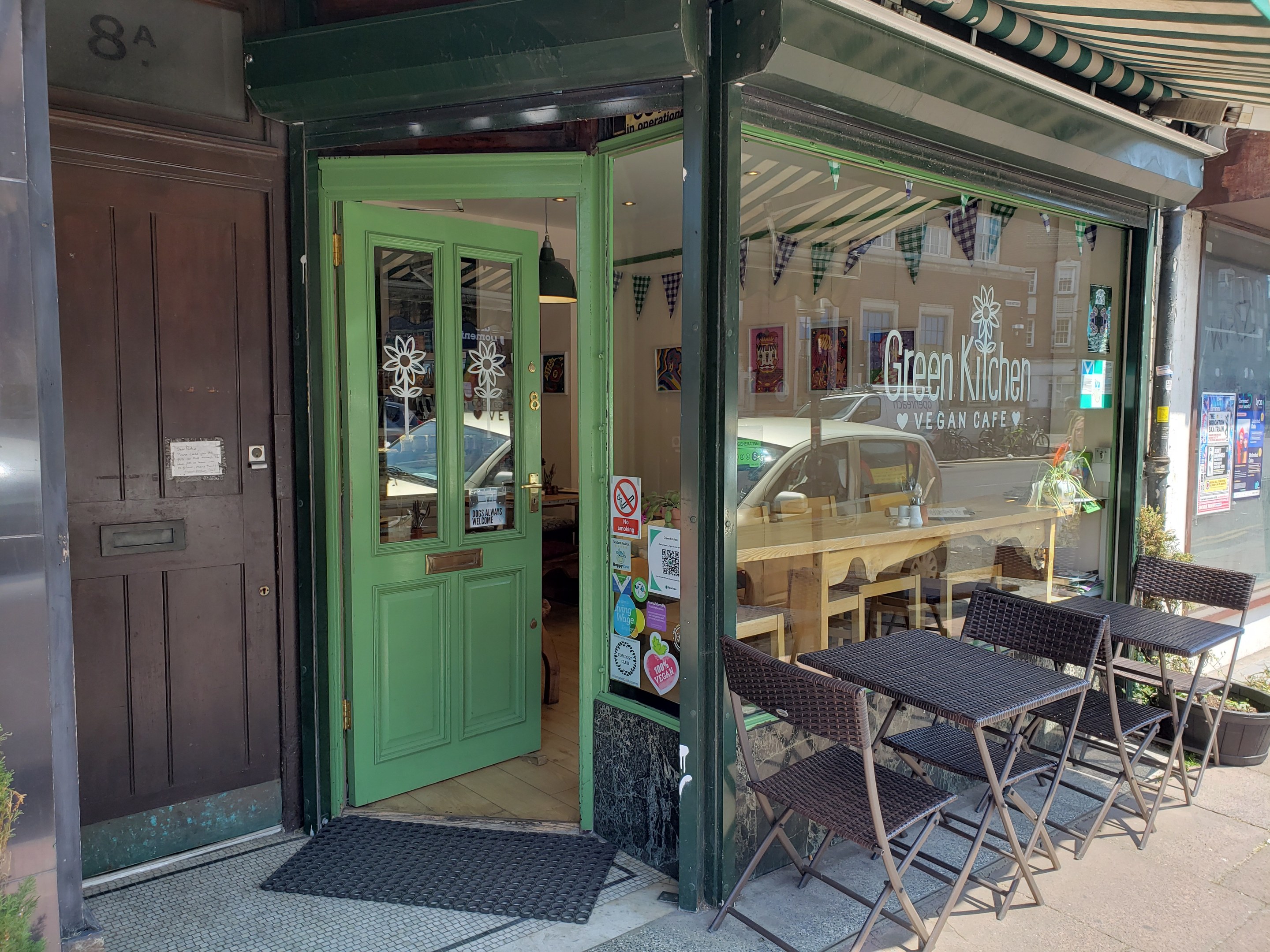 Außensitzbereich des Green Kitchen Vegan Cafe mit Tischen, Stühlen, Pflanzen, einer Matte und einer Glastür mit Postern, die in einen Innenraum mit Tischen, Stühlen und einer Hängelampe führen.