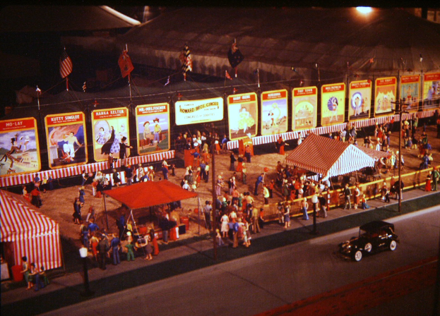 Ein altes Nacht-Karnevalsbild mit Menschen, einem Auto, Masten mit Fahnen, einem Zaun, einem Zelt, Brettern mit Bildern und Text sowie einer Straßenlaterne.