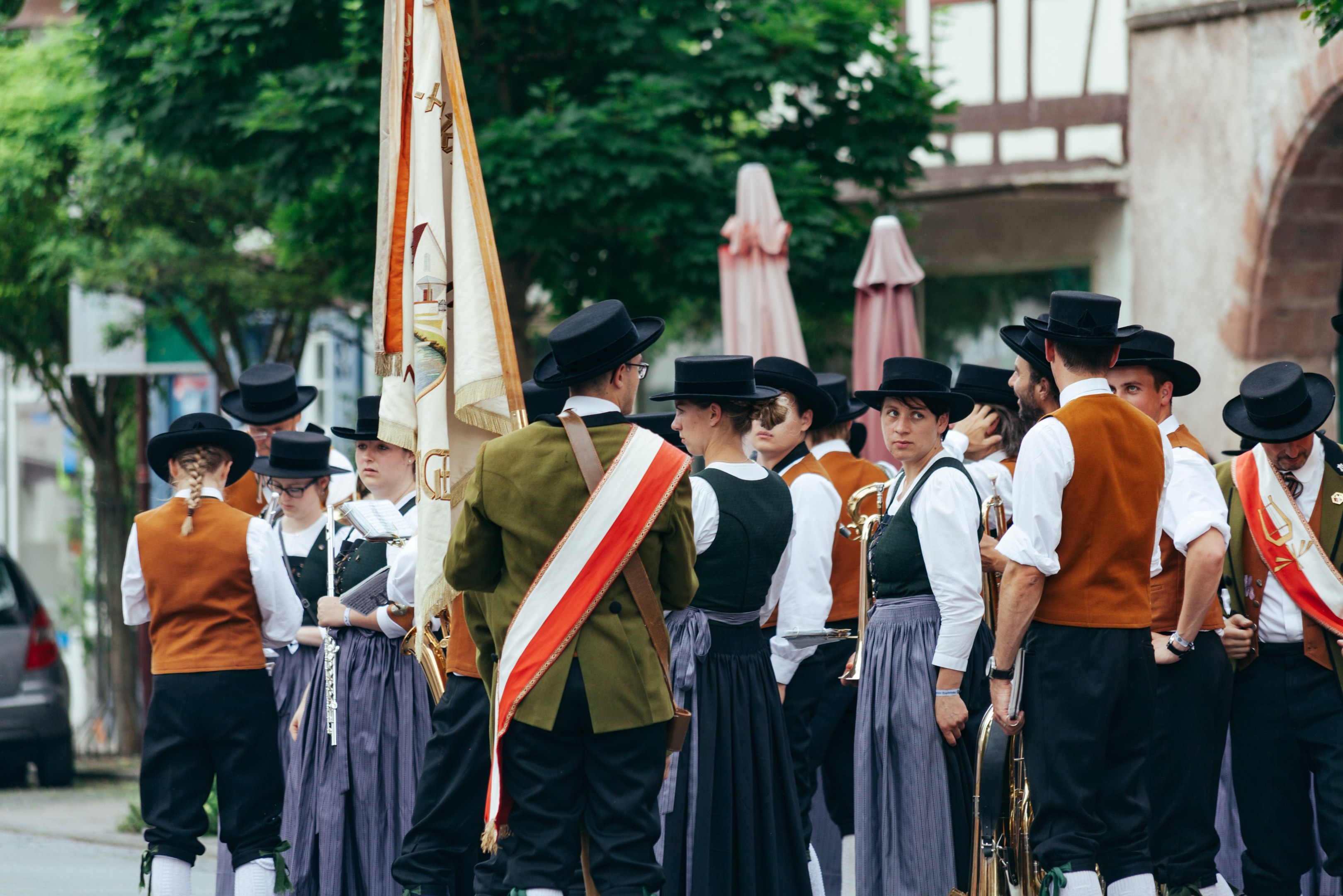 Gruppe von Menschen in traditioneller bayrischer Tracht, die eine Straße entlangmarschieren, einige halten Musikinstrumente und Fahnen, im Hintergrund sind Bäume, Gebäude und ein Auto zu sehen.