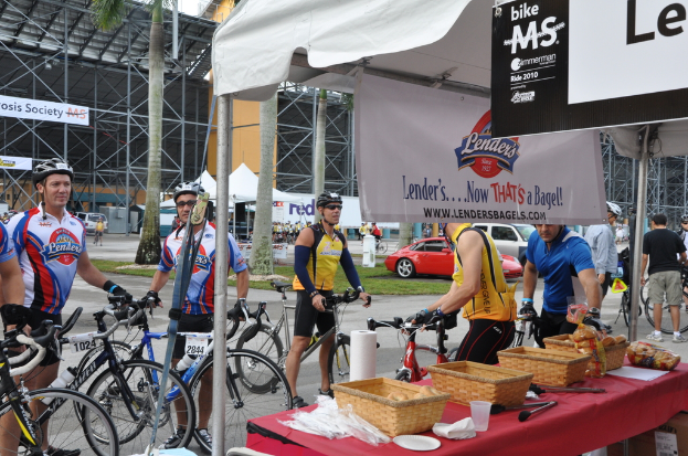 Eine Straße mit einigen Menschen in der Nähe von Fahrrädern, ein Geschäft mit Gegenständen auf einem Tisch, Bäume, Zelte, an eine Hütte angebrachte Banner, Fahrzeuge und Gras.