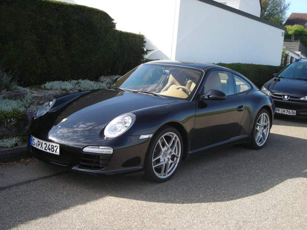 Zwei schwarze Porsche 911 vor einem weißen Haus geparkt, umgeben von üppigen grünen Pflanzen und Bäumen, mit einem hellblauen Himmel im Hintergrund.