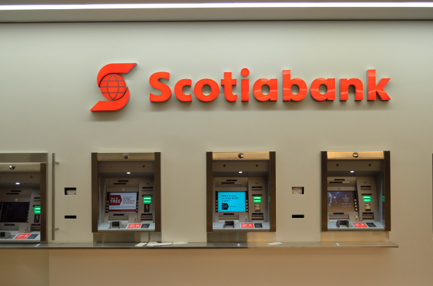 Ein modernes Scotiabank-Geldautomatengerät mit schwarzer und silberner Optik, einem großen Display und Bedienungstasten, vor einer hellgrauen Wand mit gerahmten Bildern und der Aufschrift "Scottish Bank" im Hintergrund.