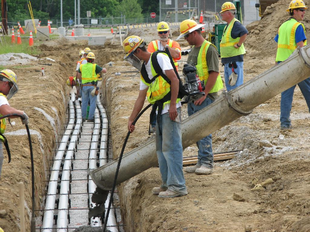 Eine Baustelle mit Arbeitern in Jacken und Helmen, Absperrgittern, Zaun, Bäumen, Pfählen, Erde und Rohren im Hintergrund.