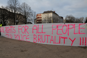 Eine Gruppe von Menschen, die auf dem Boden steht und eine Fahne hält, auf der 'Rechte für alle Menschen Stoppt Polizeigewalt' steht, mit einem Straßenschild, einem Schild, Bäumen, Gebäuden mit Fenstern und einem bewölkten Himmel im Hintergrund.