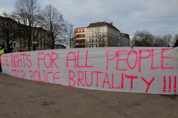 Eine Gruppe von Menschen, die auf dem Boden steht und eine Fahne hält, auf der 'Rechte für alle Menschen Stoppt Polizeigewalt' steht, mit einem Straßenschild, einem Schild, Bäumen, Gebäuden mit Fenstern und einem bewölkten Himmel im Hintergrund.