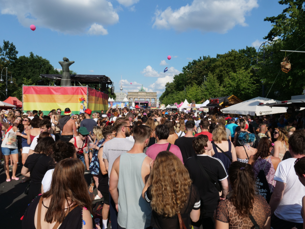 Eine große Menschenmenge, die eine Straße mit Zelten, Bäumen, Pfählen, Laternen und einer Statue säumt, mit Gebäuden im Hintergrund und einem bewölkten Himmel mit Ballons bei der Christopher Street Day Parade in Berlin.