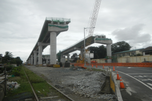 Baustelle mit Brücke, Straße mit Verkehrskegeln, Bahnschiene, Steine, Gras, Bäume, Gebäude und bewölkter Himmel.