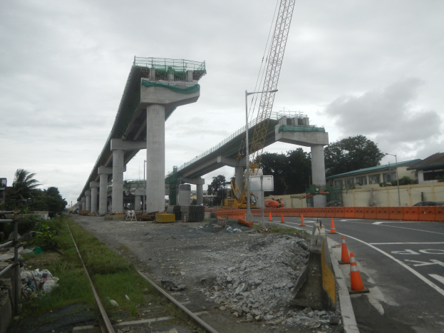 Baustelle mit Brücke, Straße mit Verkehrskegeln, Bahnschiene, Steine, Gras, Bäume, Gebäude und bewölkter Himmel.