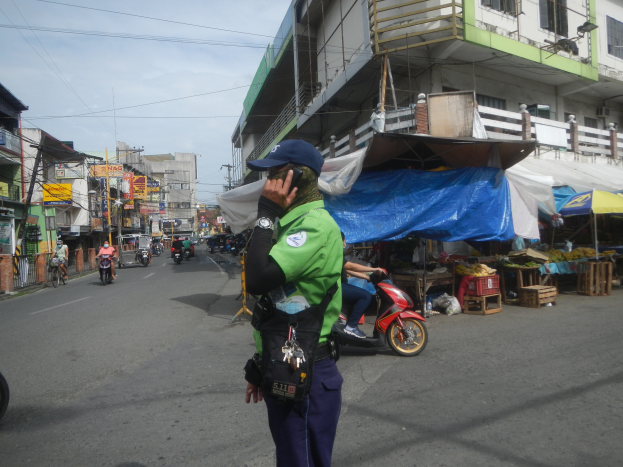 Ein Polizeibeamter in grüner und blauer Uniform und einer blauen Mütze steht an der Seite einer Straße und spricht auf einem Handy, während hinter ihm Fahrzeuge auf der Straße fahren, Stände mit verschiedenen Gegenständen zu seiner Rechten und ein bewölkter Himmel mit einem Gebäude mit Fenstern im Hintergrund.