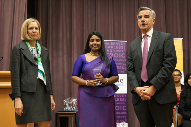 Drei Personen stehen auf einer Bühne bei den Women's Leadership Awards 2016: eine Frau in einem lila Kleid mit einem Preis in der Hand, flankiert von einem Mann in einem schwarzen Blazer, weißem Hemd und pinker Krawatte, mit Preisen auf einem Tisch und einem Podium links daneben.