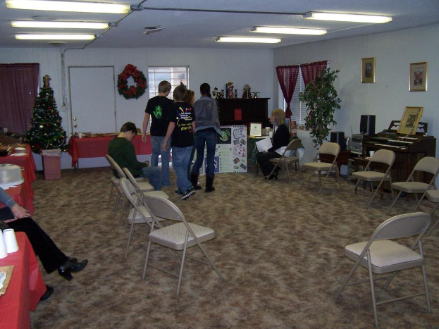 Ein Raum mit mehreren Stühlen, Tischen und Personen, einige stehen, einige sitzen. Auf den Tischen stehen Tassen und Gefäße. Im Hintergrund ist eine Wand mit einem Strauß, einem Vorhang, einer Tür, Fotorahmen und Fenstern zu sehen. Ein Weihnachtsbaum und ein Piano sind zu erkennen, mit Röhrenleuchten an der Decke.