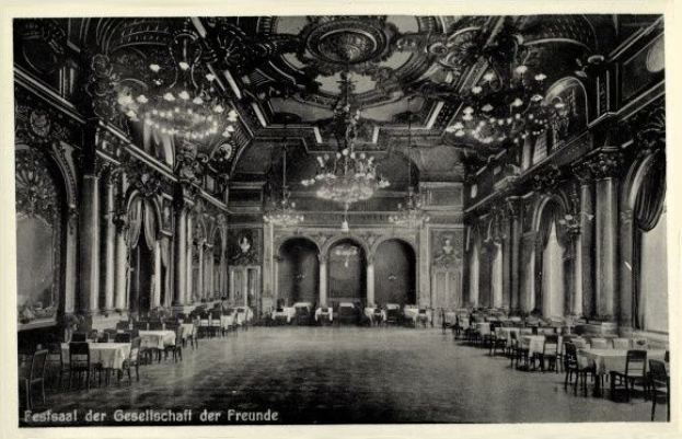 Schwarz-weiß-Foto eines großen Raums mit Tischen, Stühlen, Säulen, Bögen und Kronleuchtern, beschriftet mit 'Festival der Gesellschaft der Freunde, Berlin' unten.
