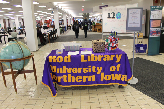 Eine Ausstellung mit einem Tisch, auf dem verschiedene Landesflaggen, eine Box, Papiere und ein Banner liegen, eine Kugel links daneben, ein Anschlagbrett rechts mit einigen Menschen in der Nähe und mehreren Tischen und Stühlen links.