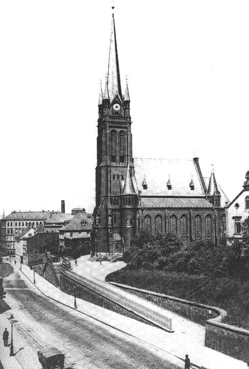 Schwarzes und weißes Foto einer Stadtstraße mit der St. Peter Kirche in Wiesbaden im Hintergrund, flankiert von Gebäuden, Bäumen, Fahrzeugen und Passanten.