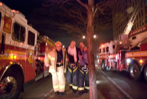 Drei Personen vor einem Löschfahrzeug auf der linken Seite und einem Gebäude auf der rechten Seite mit einem Baum in der Mitte.