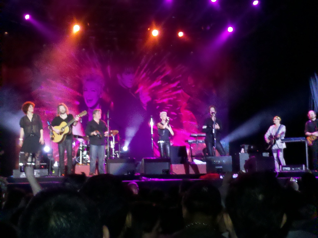 Eine Bühne mit Musikern, die Gitarren spielen und in Mikrofone singen, begleitet von Trommeln, mit einem Publikum davor und Lichtern und einem Bildschirm im Hintergrund.