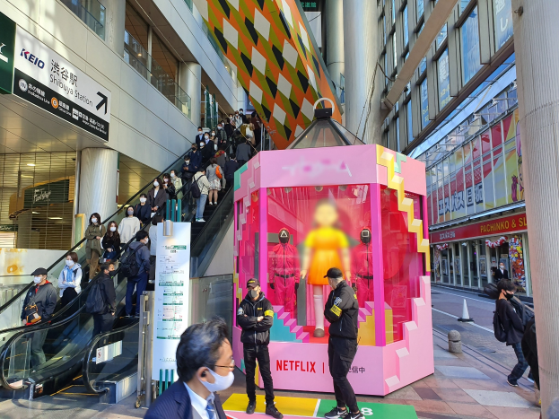 Eine Gruppe von Menschen steht vor einem gläsernen Gebäude mit Säulen und Texttafeln, einige tragen Masken, während eine Rolltreppe mit Menschen in der Nähe nach oben führt; eine pinkfarbene Box in der Mitte zeigt Schaufensterpuppen und trägt die Aufschrift "Netflix's new Netflix booth in Tokyo."