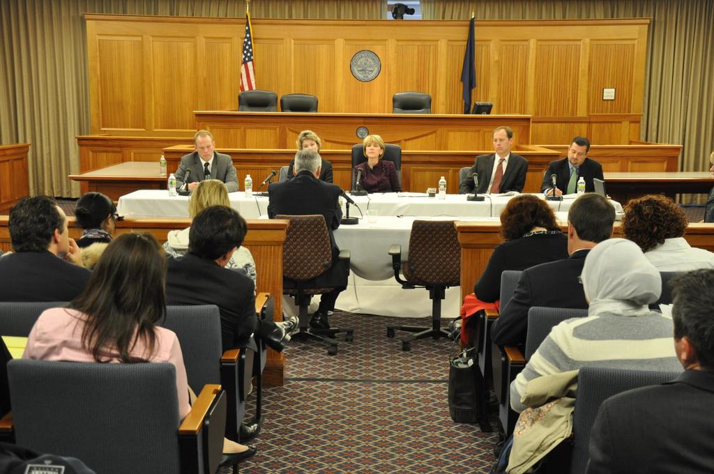 Ein Versammlungsraum mit einer Gruppe von Menschen, die auf Stühlen sitzen, zwei ausländischen Flaggen, einem orangefarbenen Schreibtisch mit Wasserflaschen und einem Mikrofon.