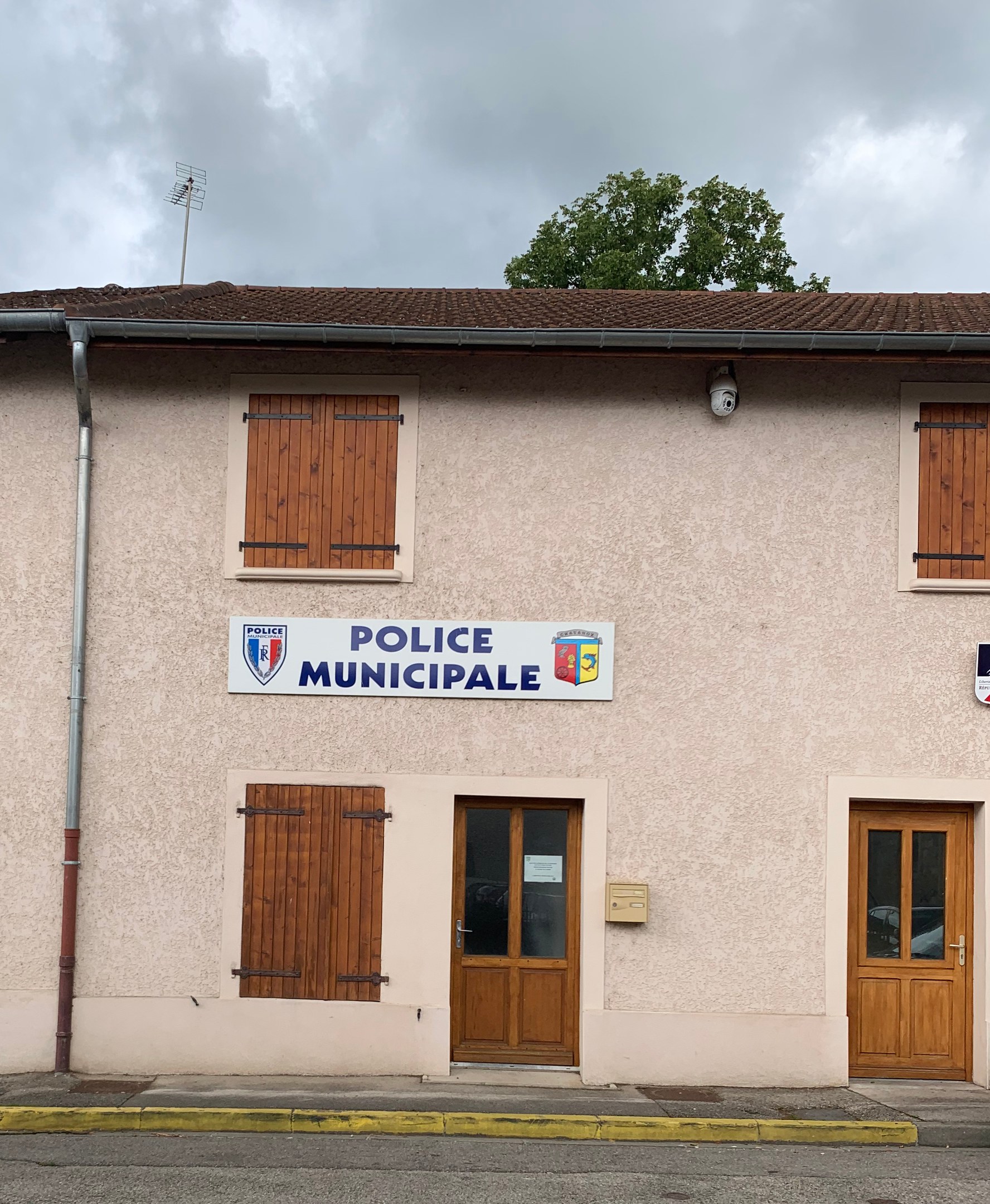 Polizeigebäude mit einem Schild, Fenstern, Türen, einer Namensplakette, einer Leitung, einem Baum und einem bewölkten Himmel im Hintergrund.