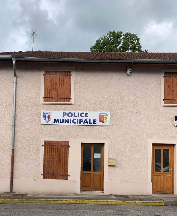 Polizeigebäude mit einem Schild, Fenstern, Türen, einer Namensplakette, einer Leitung, einem Baum und einem bewölkten Himmel im Hintergrund.