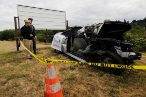 Ein Polizist in Uniform steht auf dem Gras neben einem schwer beschädigten Auto mit eingedellter Motorhaube und zersplitterten Fenstern, mit einem Verkehrskegel und einem Band an einem Pfahl in der Nähe und Bäumen und einem bewölkten Himmel im Hintergrund.