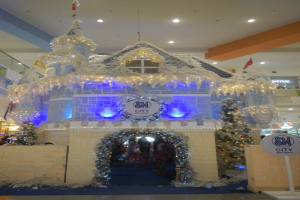Ein festliches Weihnachtsdorf mit einer burgenähnlichen Struktur, die mit Lichtern geschmückt ist, umgeben von Menschen in einem Einkaufszentrum mit beleuchteter Decke und einer Tafel auf der rechten Seite.
