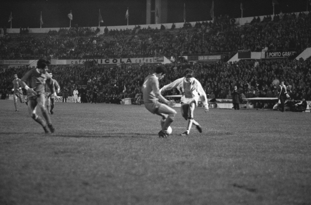 Schwarzes und weißes Bild von Männern, die Fußball spielen, in einem Stadion mit Zuschauern auf den Tribünen und Fahnen im Hintergrund.