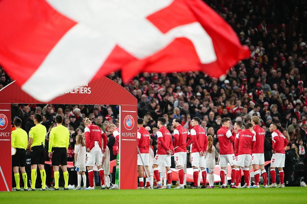 Eine Gruppe von Menschen auf einem Fußballfeld mit einer roten und weißen Flagge im Vordergrund, ein Bogen mit der Aufschrift "Bayern München vs. Bayern München Wetten & Vorschau" im Hintergrund und eine große Menge im Stadion.