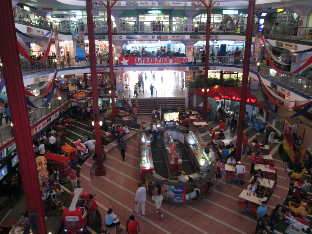 Eine belebte Einkaufszentrumszene mit Säulen, Menschen, Tischen, Fliesen, Fahnen, Geschäften, einem Banner, Treppen und einem Namensschild.