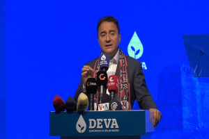 Ein Mann in Mantel und Schal steht an einem Podium mit Mikrofonen, ein Schild mit der Aufschrift "deva-Geschäftsführer sagt, es sei keine gute Idee für die Zukunft", ein Objekt und ein Bildschirm im Hintergrund.