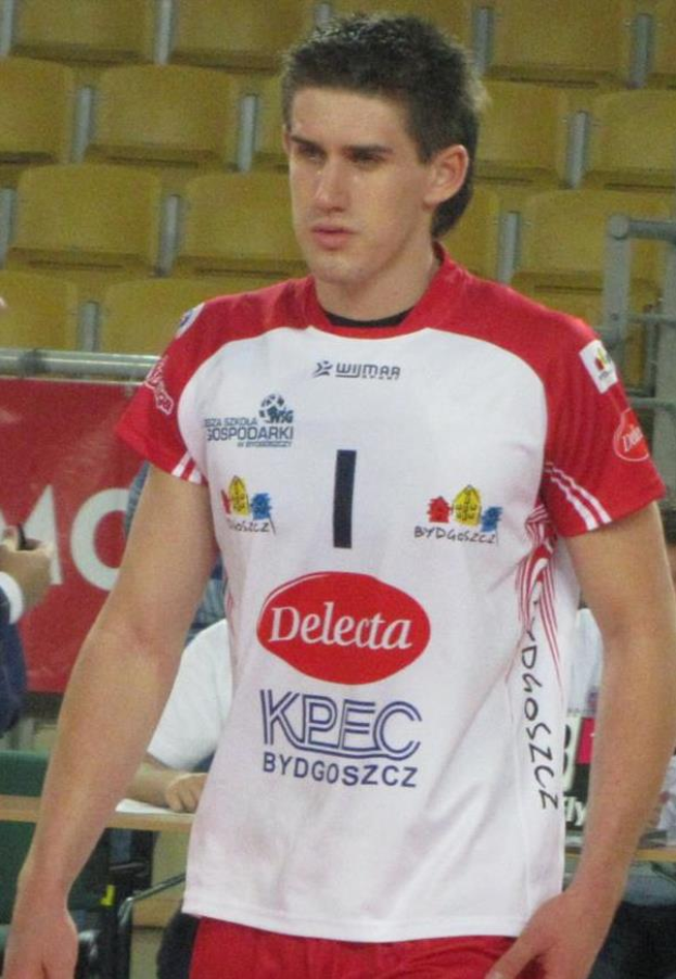 Ein Mann in einem roten und weißen Handballtrikot steht auf einem Feld, umgeben von Zuschauern, mit einem roten Banner im Hintergrund, auf dem "Krasnoyarsk vs Bydgoszcz" steht und sieht entschlossen aus.