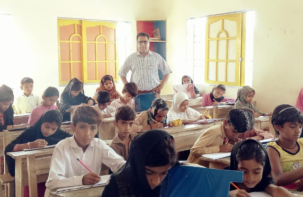 Eine Gruppe von Kindern sitzt an Tischen in einem Klassenzimmer in Pakistan, mit Büchern und Stiften in den Händen, mit einer Person im Hintergrund und einem Bücherregal im Hintergrund und Fenstern an den Wänden.