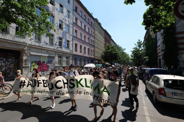 Eine Gruppe von Menschen marschiert mit einem Banner die Straße entlang, mit parkenden Autos auf der rechten Seite, einem Fahrradfahrer auf der linken Seite, Gebäuden, Bäumen und einem Schild im Hintergrund, unter einem sichtbaren Himmel.