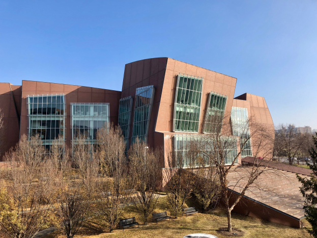Großes mehrstöckiges Gebäude mit zahlreichen Fenstern, umgeben von Bäumen, Bänken, Gras und einem Laternenmast, mit anderen Gebäuden und einem klaren blauen Himmel im Hintergrund, als der neue Campus der University of Illinois at Chicago identifiziert.