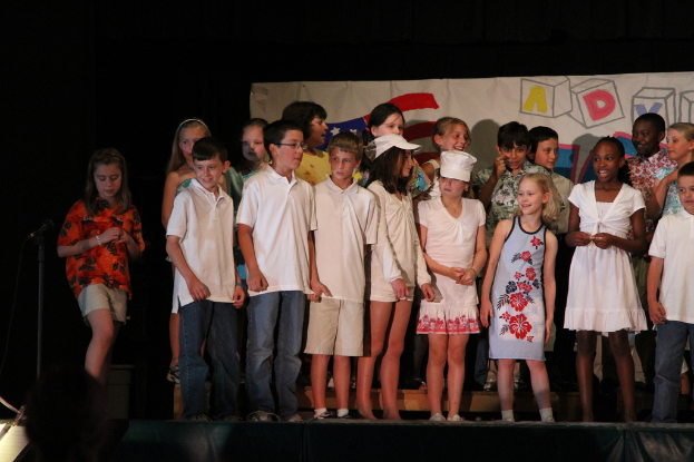 Mehrere Kinder stehen auf einer Bühne, wobei zwei in der Mitte Mützen tragen, ein Mikrofonständer links und ein Banner im Hintergrund.