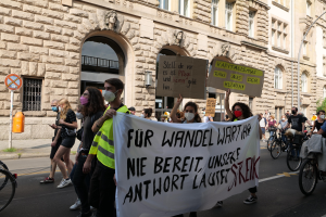 Eine Gruppe von Menschen marschiert bei einer Demonstration auf einer Straße, einige halten Schilder und andere fahren Fahrräder, im Hintergrund ein Gebäude mit Fenstern, Bögen, Säulen und Skulpturen.