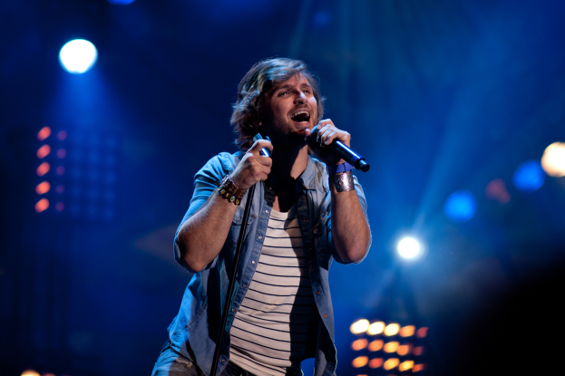 Ein Mann steht auf einer hell erleuchteten B├╝hne und singt in ein Mikrofon, eine Gitarre ├╝ber der Schulter, in einem schwarzen T-Shirt und Jeans, mit stylischem, unordentlichem Haar und einem selbstbewussten Ausdruck.