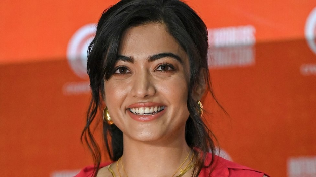 Eine Frau in einem roten Kleid mit einem eleganten Dutt und dezentem Make-up posiert strahlend, trägt schwarze High Heels und hat einen hellgelben Banner mit der Aufschrift 'Sonam Kapoors beste Schönheits Looks vom Cannes Film Festival' im Hintergrund.
