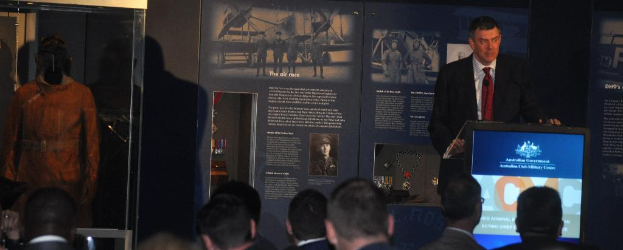 Eine Person steht an einem Rednerpult mit einer Gruppe von Menschen davor, eine Schachtel mit einer Puppe darin links daneben und Tafeln im Hintergrund mit Text, Medaillen und Bildern.