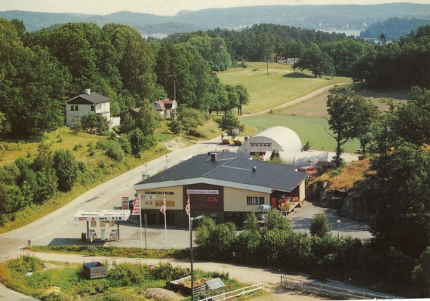 Ein altes Foto einer Tankstelle in einem Feld umgeben von Häusern, Bäumen, Pflanzen und diversen Gegenständen, mit Hügeln und einem klaren blauen Himmel im Hintergrund und Text unten.