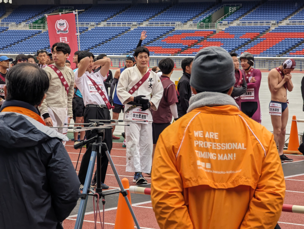 Eine Gruppe von Menschen, die auf einer Bahn mit einer Kamera auf einem Stativ in der Vordergrund, Stühle, ein Banner und Lichter im Hintergrund stehen.